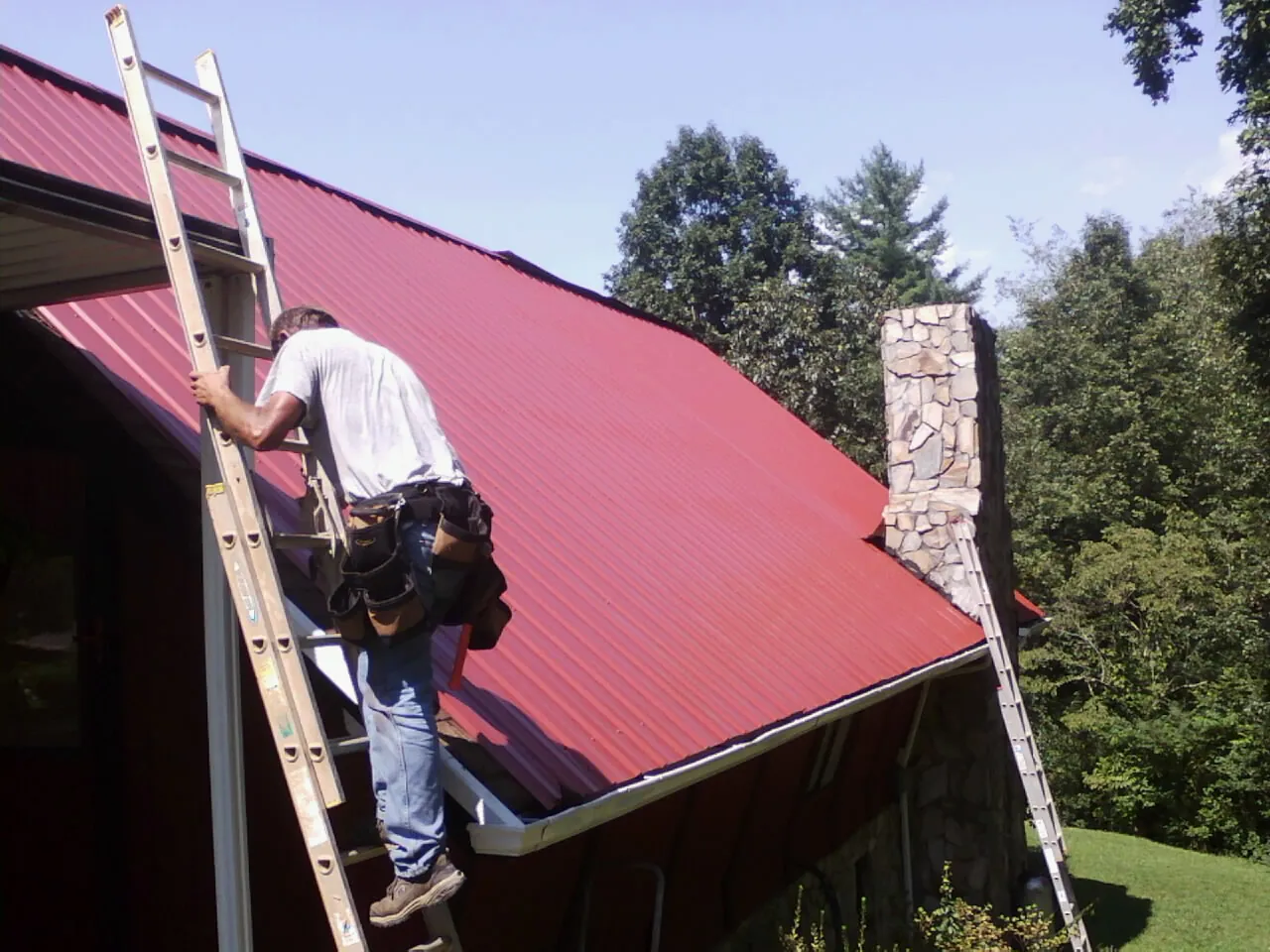 Roofer on a ladder beside a red metal roof and stone chimney — Soffit & Fascia Repair by Roofing Brothers in Boligee